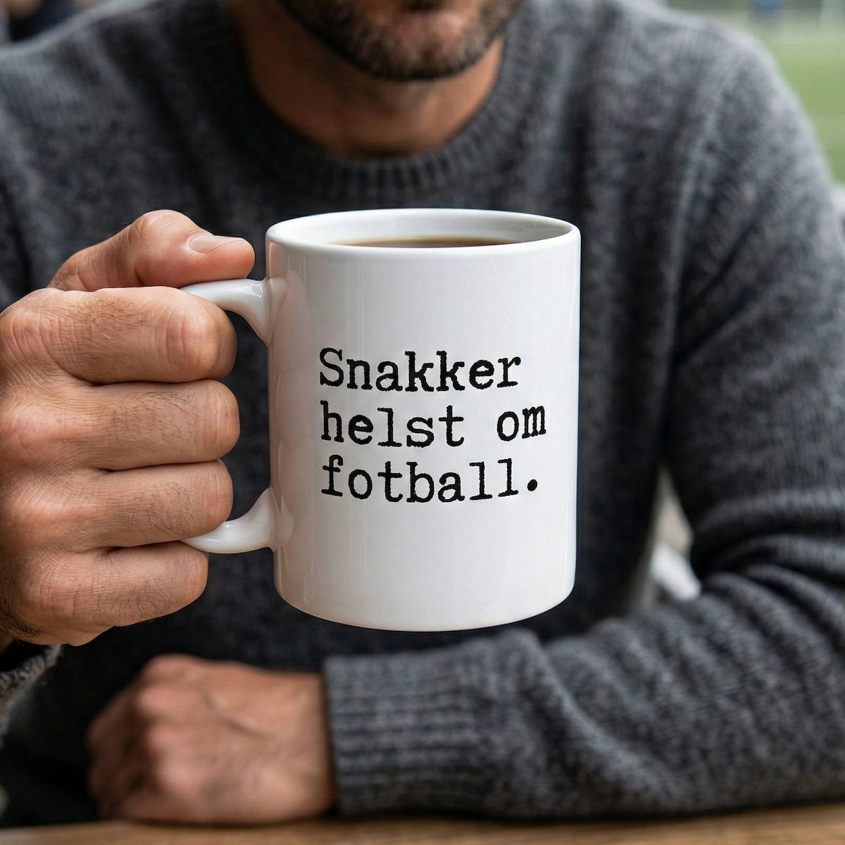 En person som holder en hvit kopp med teksten "Snakker helst om fotball" i en lun og hverdagslig setting.