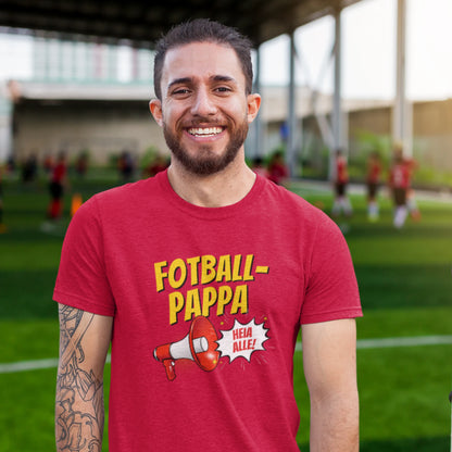 Smilende mann i rød T-skjorte med teksten "Fotball-Pappa" og en megafon, står på en fotballbane med barn som spiller i bakgrunnen.
