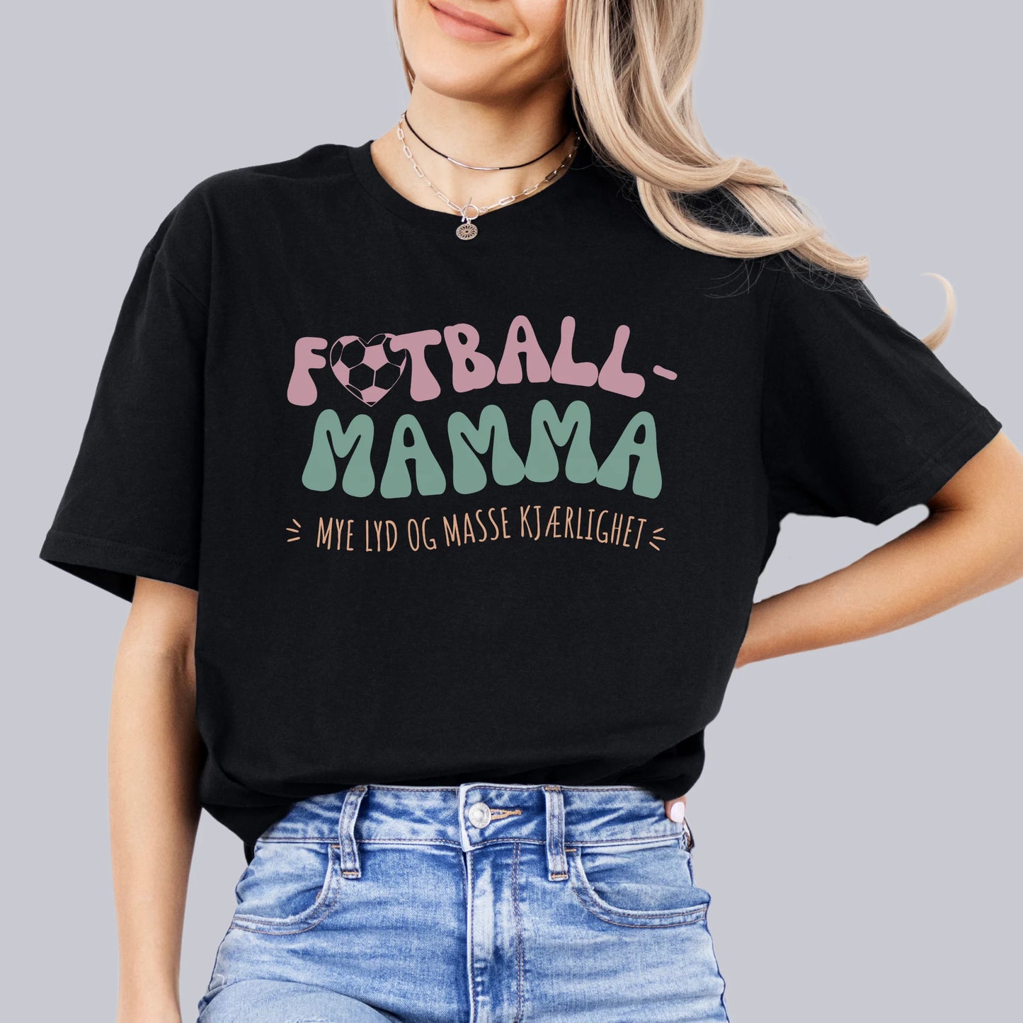 Kvinne i svart T-skjorte med teksten "Fotball-Mamma – Mye lyd og masse kjærlighet" i en fargerik, leken stil.






