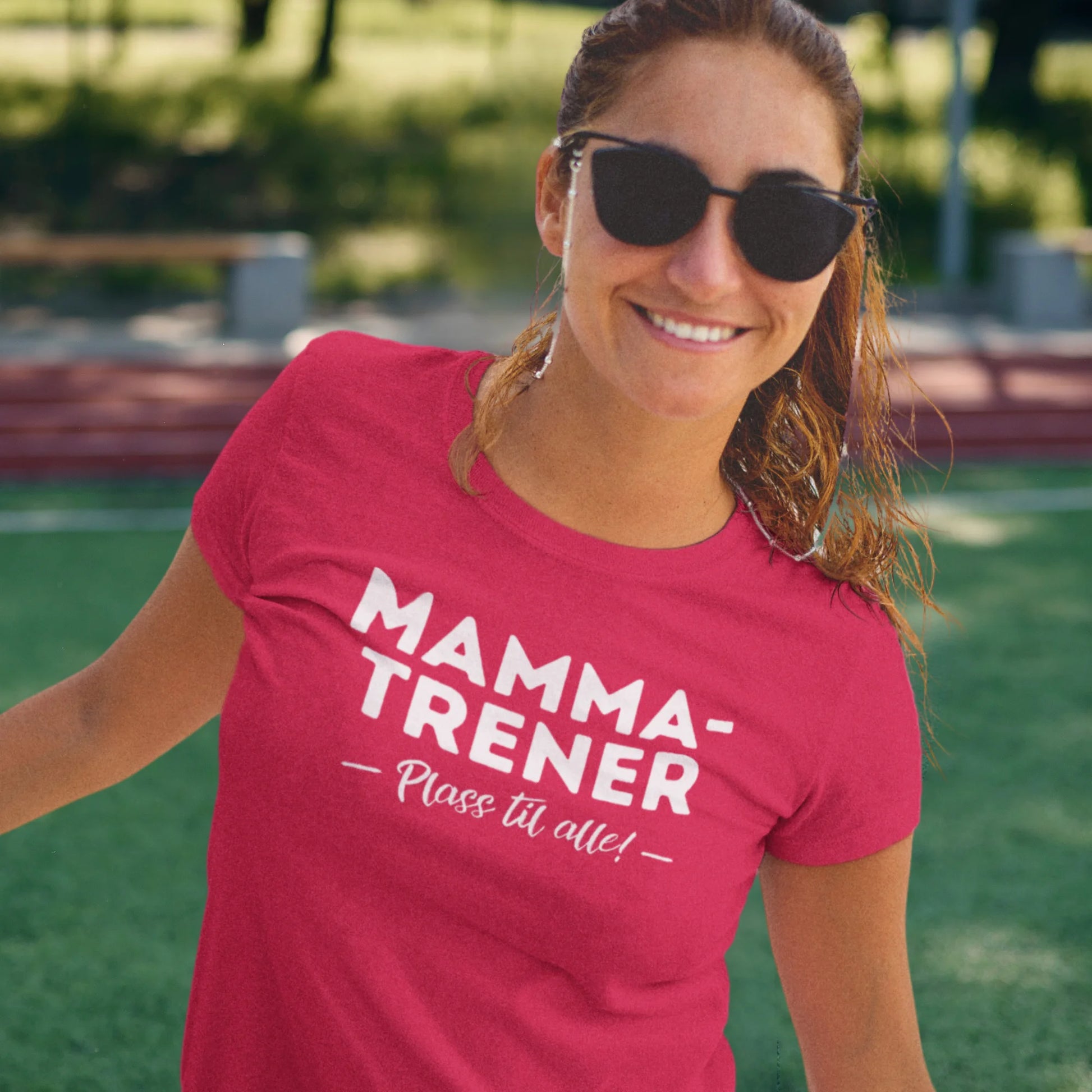 Smilende kvinne i solbriller og rød T-skjorte med teksten "Mamma-Trener – Plass til alle!", står på en fotballbane i solskinn.