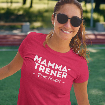 Smilende kvinne i solbriller og rød T-skjorte med teksten "Mamma-Trener – Plass til alle!", står på en fotballbane i solskinn.