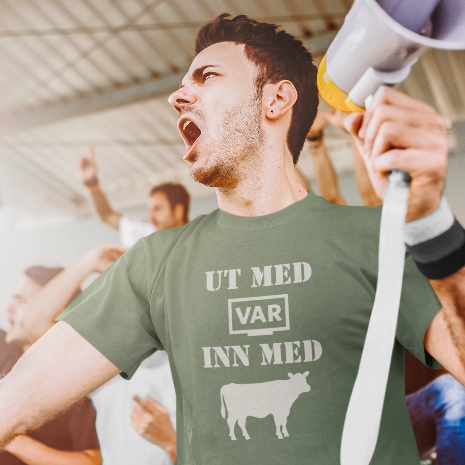 Military Green T-skjorte med teksten 'UT MED VAR, INN MED KUA', båret av en fotballsupporter.