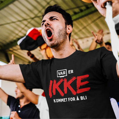Engasjert fotballsupporter roper på tribunen iført en svart T-skjorte med teksten "VAR er IKKE kommet for å bli" i hvitt og rødt. Bakgrunnen viser andre supportere som heier.