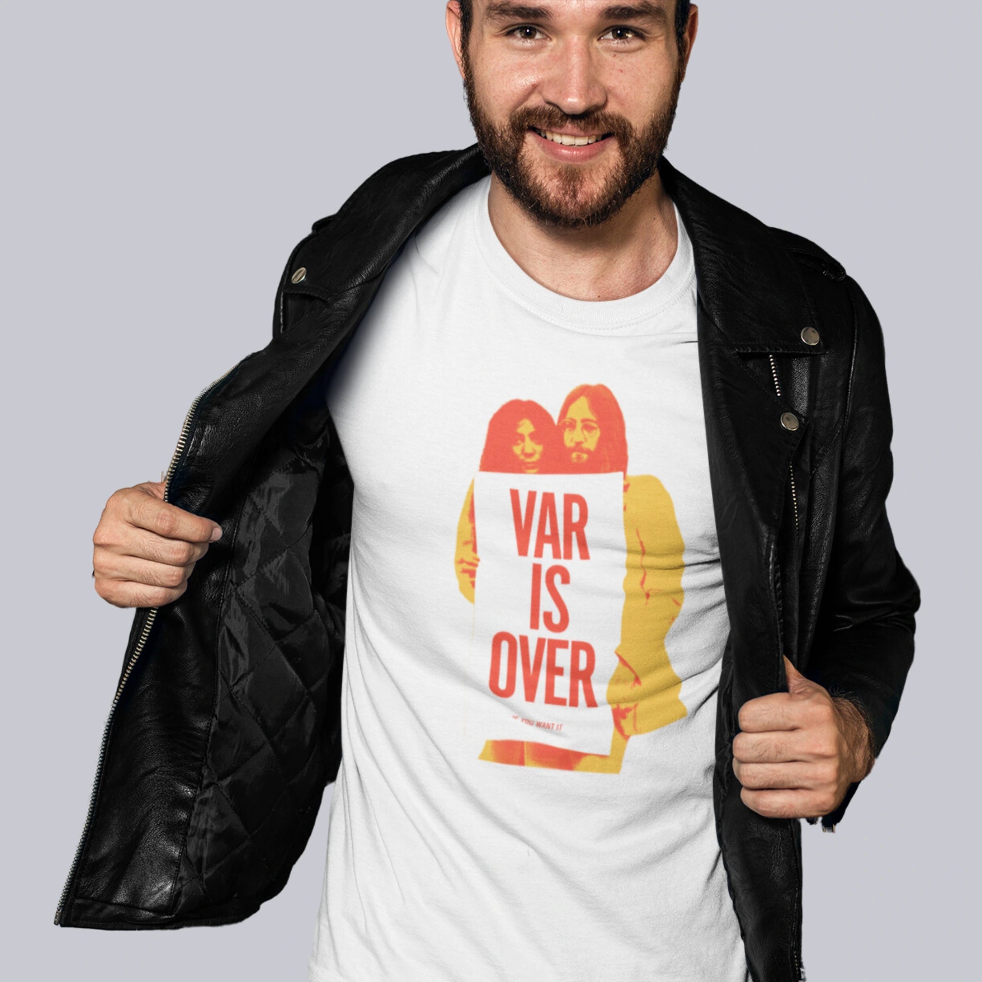 Smilende mann i skinnjakke viser frem en hvit T-skjorte med trykket "VAR IS OVER" inspirert av den ikoniske John Lennon-plakaten. Han står foran en vegg med graffiti og slitt maling.