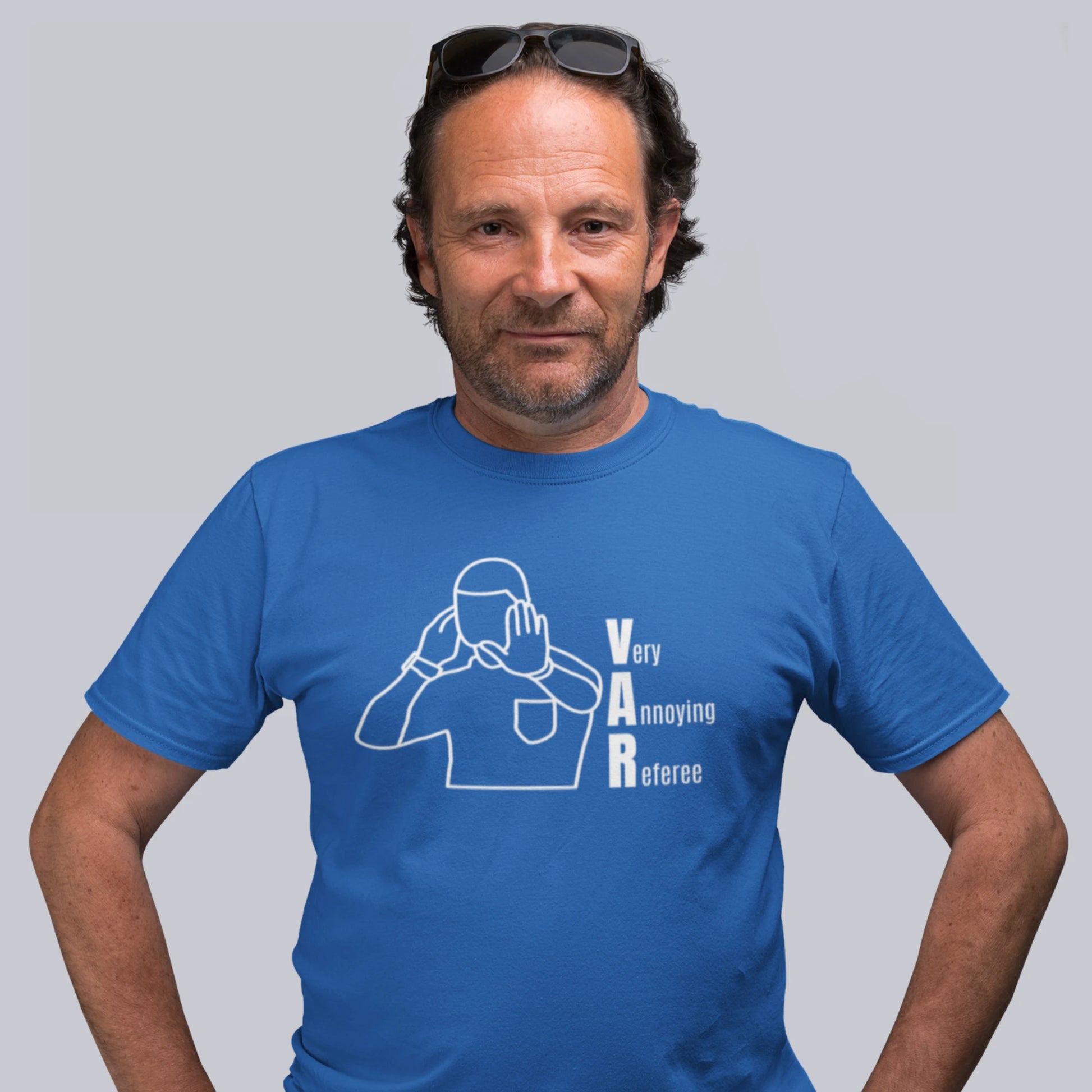 mann i blå T-skjorte med motiv av en dommer som signaliserer VAR, og teksten "Very Annoying Referee" plassert vertikalt ved siden av figuren. Han har solbriller på hodet og står mot en nøytral bakgrunn.